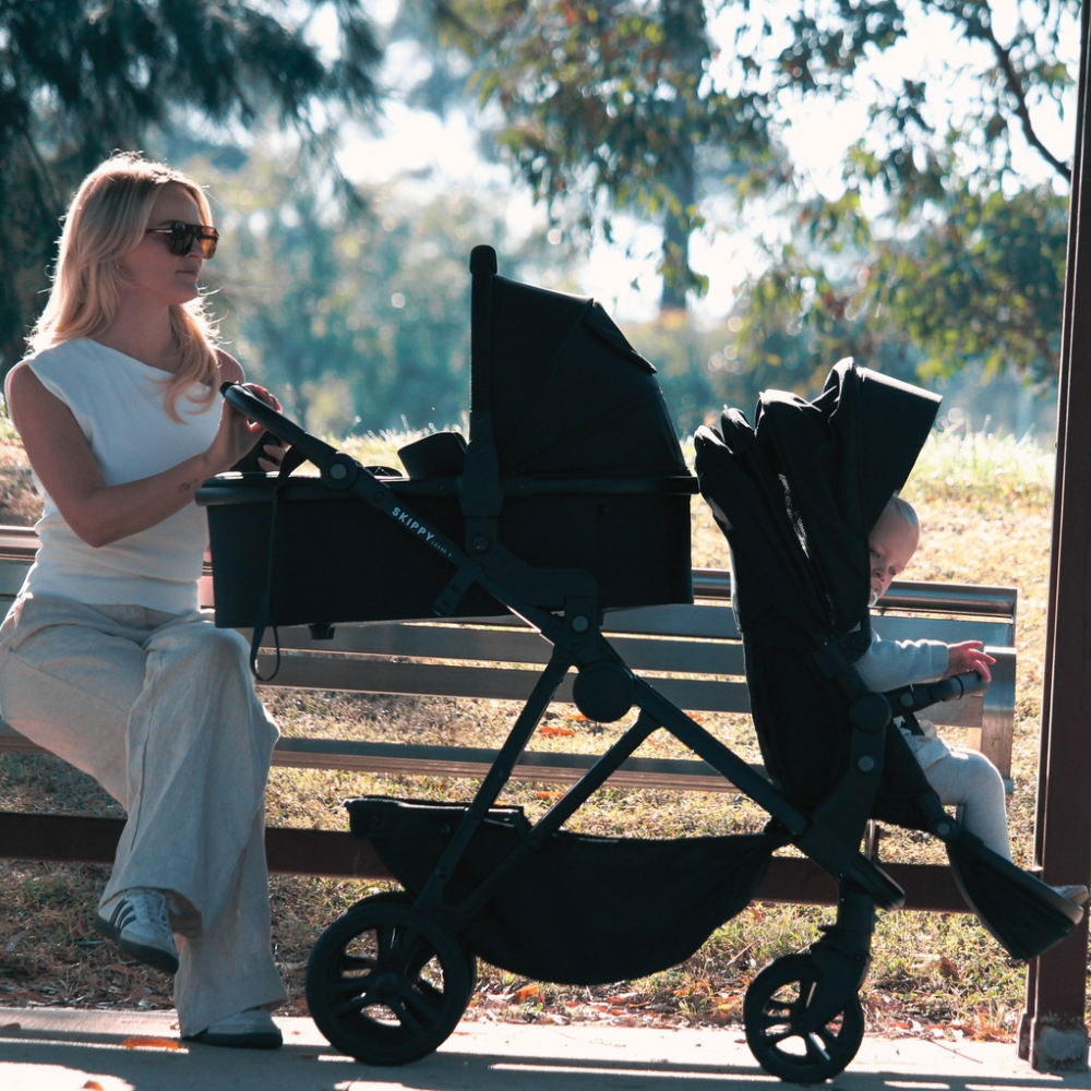 Mother sitting beside Lusso Gen 3 double pram with bassinet and toddler seat in park
